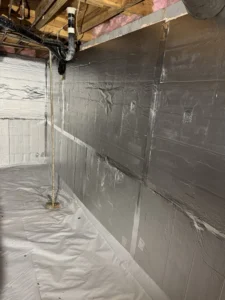 A section of a crawlspace with silver insulation panels installed on the walls, sealed tightly, and a white vapor barrier on the floor, with wooden beams and plumbing visible overhead.