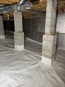 A fully encapsulated crawlspace with a white vapor barrier covering the floor, sealed around concrete support pillars, and silver insulation on the walls, with HVAC ducts and wiring visible above.