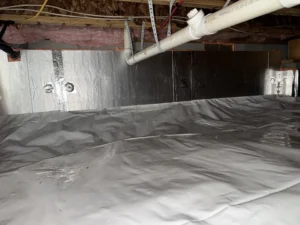 A fully encapsulated crawlspace with a reflective plastic liner covering the ground, white PVC pipes, electrical wiring, and a dehumidifier unit installed, showcasing a dry and well-maintained space.