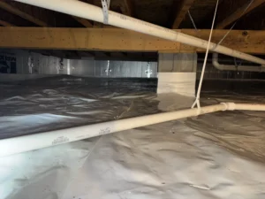 A fully encapsulated crawlspace with a reflective plastic vapor barrier covering the ground, white PVC pipes hanging from wooden beams, and a clean, dry environment.